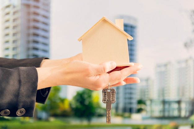 close-up-female-s-hand-holding-wooden-house-model-keys-against-blurred-building-backdrop_23-2148038696.jpg