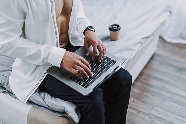 Focus on top view of guy wearing unbuttoned classic shirt and sitting on bed. He is having cup of coffee and holding notebook on knees while typing on it-ID：1431995651.jpg